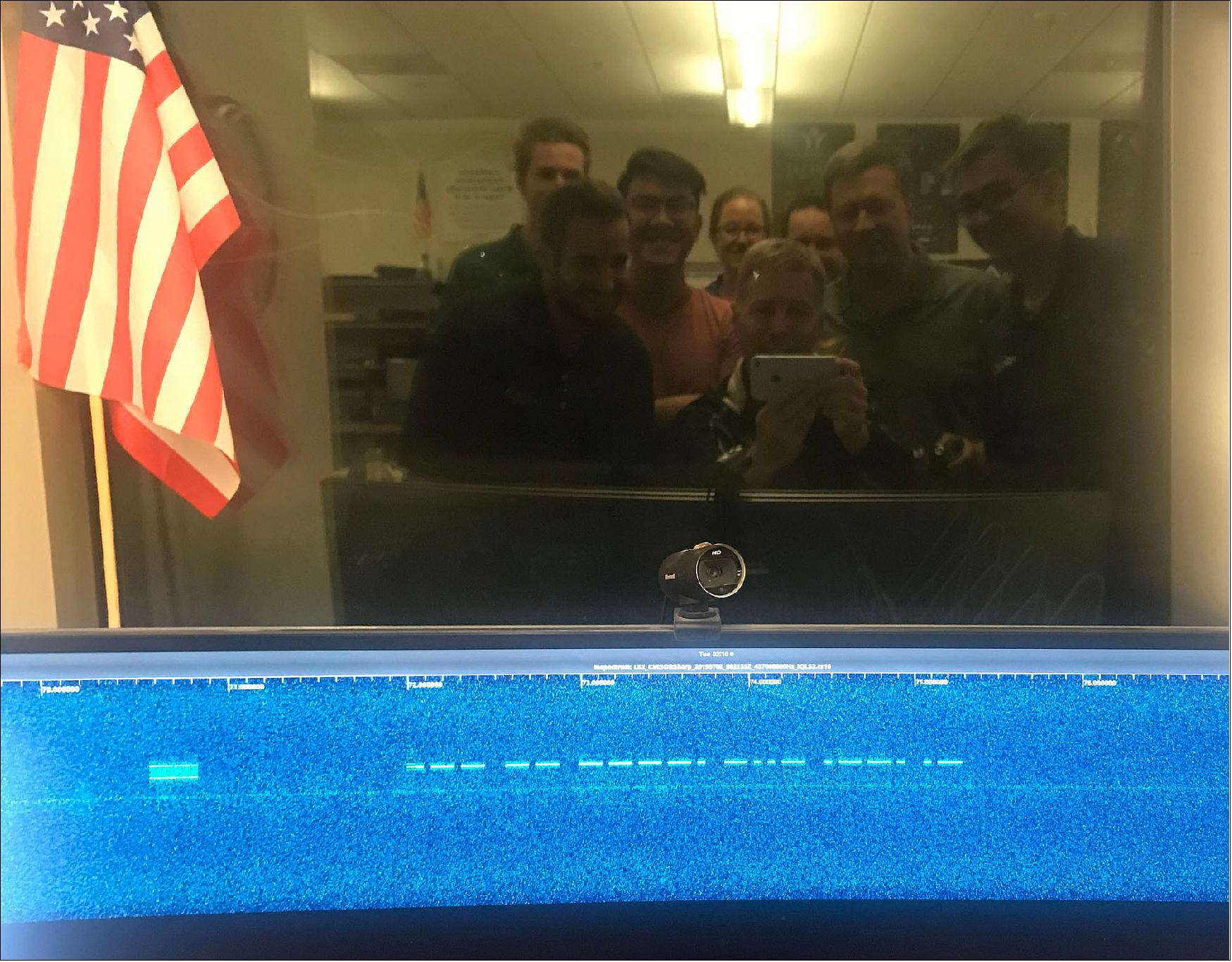 Figure 17: LightSail-2 Morse code selfie. Members of the LightSail-2 mission team can be seen in a reflection above a display showing the spacecraft’s Morse code beacon after it was first detected on 2 July 2019 at Cal Poly San Luis Obispo in California (image credit: Dave Spencer, The Planetary Society)