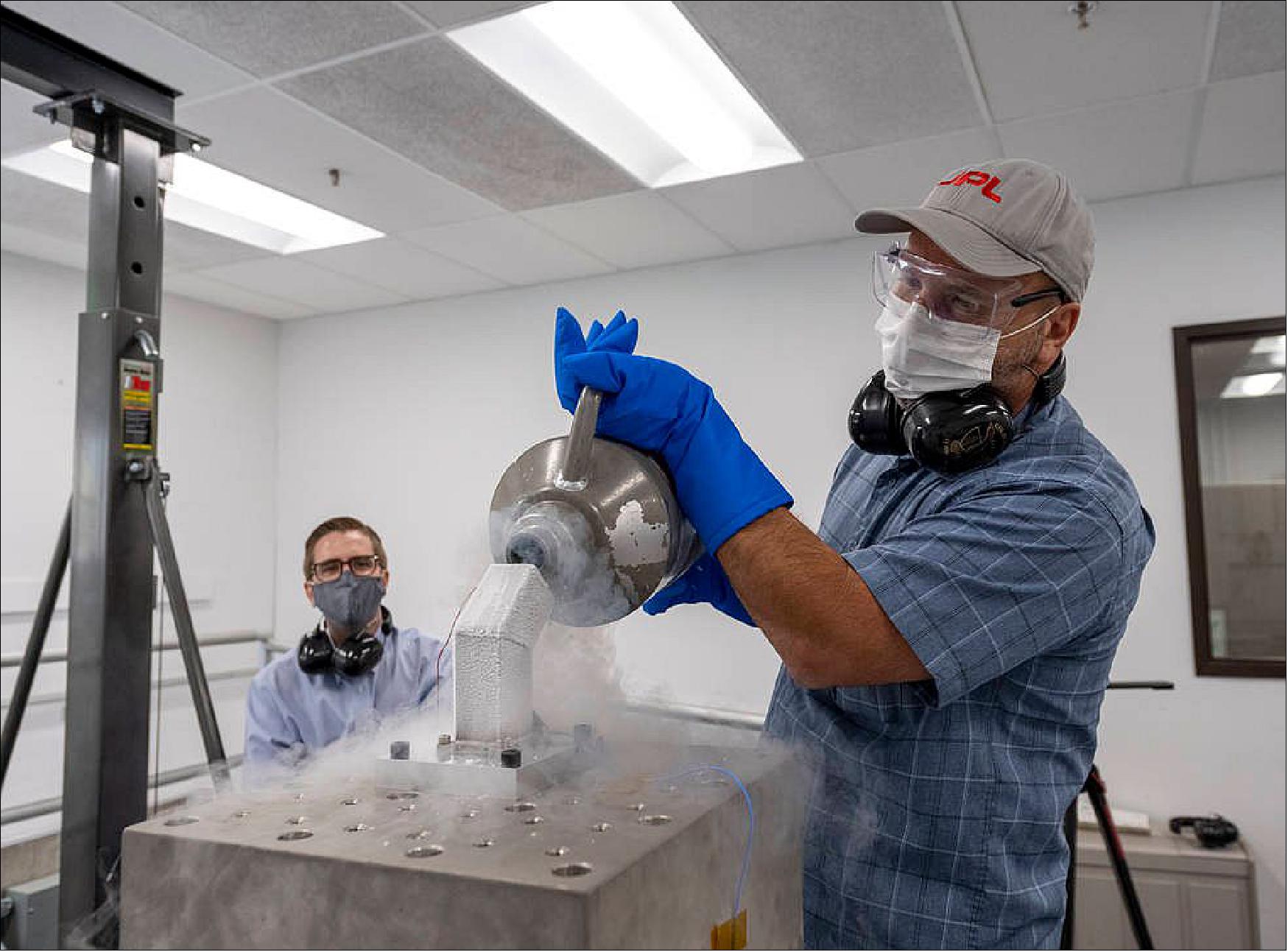 Figure 36: Andrew Kennett (left) watches as Dominic Aldi (right) uses liquid nitrogen to cool a motor integrated bulk metallic glass gearbox prior to shock testing it. The motor and gearbox are inside the frosty metal “bucket” that contains the liquid nitrogen. The tooling, including the “bucket” is designed to be mounted both vertically (shown) and horizontally on the cube for testing the motor and gearbox in three orientations (image credit: NASA/JPL-Caltech)