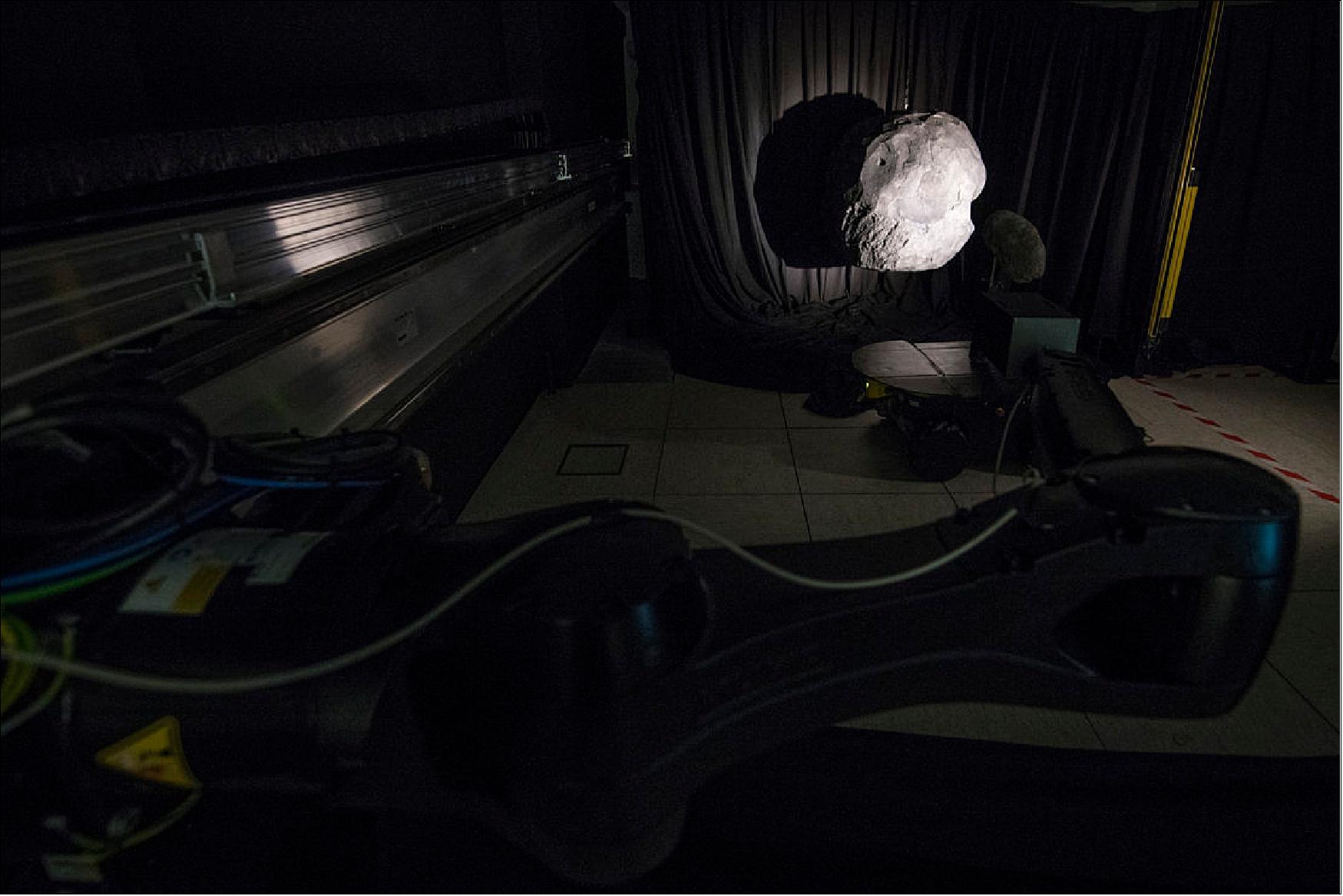 Figure 55: A camera, mounted on a robot arm that moves along a 33-m long track, approaches a pair of 3D-printed asteroid models. To simulate space, the chamber is kept dark for the testing, except for a single Sun-like light source (image credit: ESA–G. Porter, CC BY-SA 3.0 IGO)