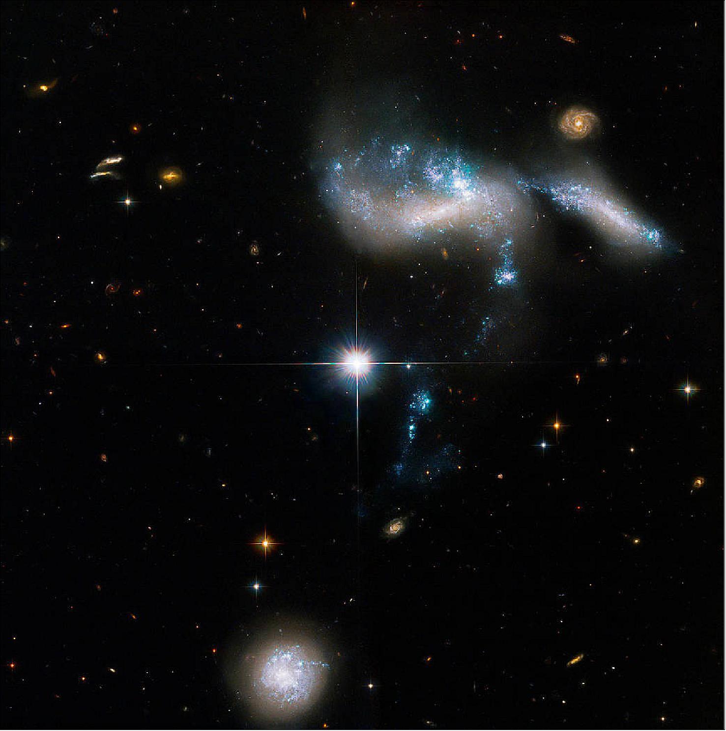 Figure 39: The bright, distorted clump of young blue-white stars (top-right of center) is NGC 1741. Although it appears to be a single galaxy, NGC 1741 is actually a pair of colliding dwarf galaxies. Another dwarf, cigar-shaped galaxy to the pair’s right joins their dance with a thin, blue stream of stars that connects the trio. HGC 31’s fourth member is revealed by a stream of young blue stars that point to the galaxy (bottom-left of center) and indicate its interaction with the other three. The bright object in the center of the image is a star situated between Earth and HCG 31 [image credit: NASA, ESA, and J. Charlton (Pennsylvania State University); Image processing: G. Kober (NASA Goddard/Catholic University of America)]