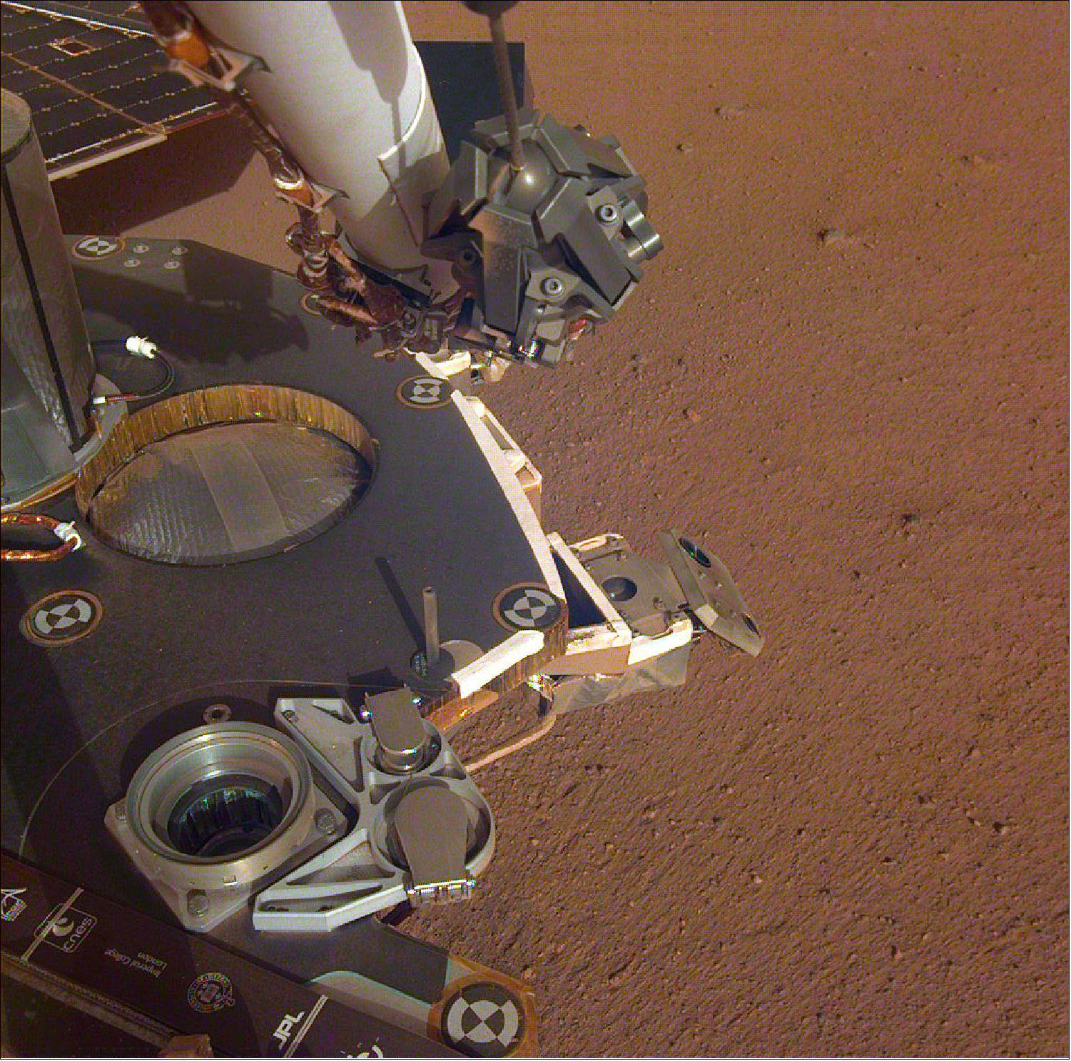 Figure 53: A partial view of the deck of NASA's InSight lander, where it stands on the Martian plains Elysium Planitia. The color-calibrated image was received on 4 December 2018 (Sol 8). InSight's robotic arm with its stowed grapple can be seen above the deck, and jutting out from the front of the deck is one of the boxy attitude control system thrusters that helped control the spacecraft's landing. The circular silver inset of the propellant tank can also be seen in the middle of the image, as well as one of the connections for the aeroshell and parachute, which looks like a cupholder in the foreground. Next to the propellant tank is the UHF antenna, which helps the lander communicate with Earth. In the background, part of one of InSight's solar panels is visible (image credit: NASA/JPL-Caltech)