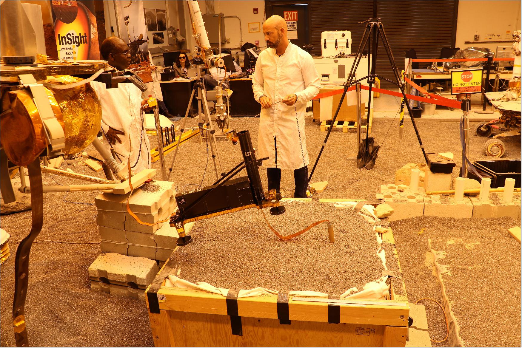 Figure 43: In this image, the model's robotic arm is lifting up part of HP3 to expose the self-hammering mole that is partially embedded in the testbed soil. Standing mid-ground are engineers Ashitey Trebi-Ollennu (left) and Troy Lee Hudson (right). Lights in the testbed intended to simulate Mars' lighting conditions give the image an orange tint. Engineers at the German Aerospace Center (DLR), which provided HP3, have also been working on strategies to help the probe (image credit: NASA/JPL, Caltech)