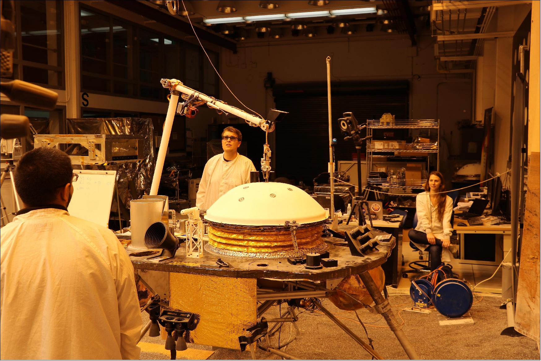 Figure 60: NASA's InSight mission tests an engineering version of the spacecraft's robotic arm in a Mars-like environment at NASA's Jet Propulsion Laboratory. The five-fingered grapple on the end of the robotic arm is lifting up the Wind and Thermal Shield, a protective covering for InSight's seismometer. The test is being conducted under reddish "Mars lighting" to simulate activities on the Red Planet (image credit: NASA/JPL-Caltech)
