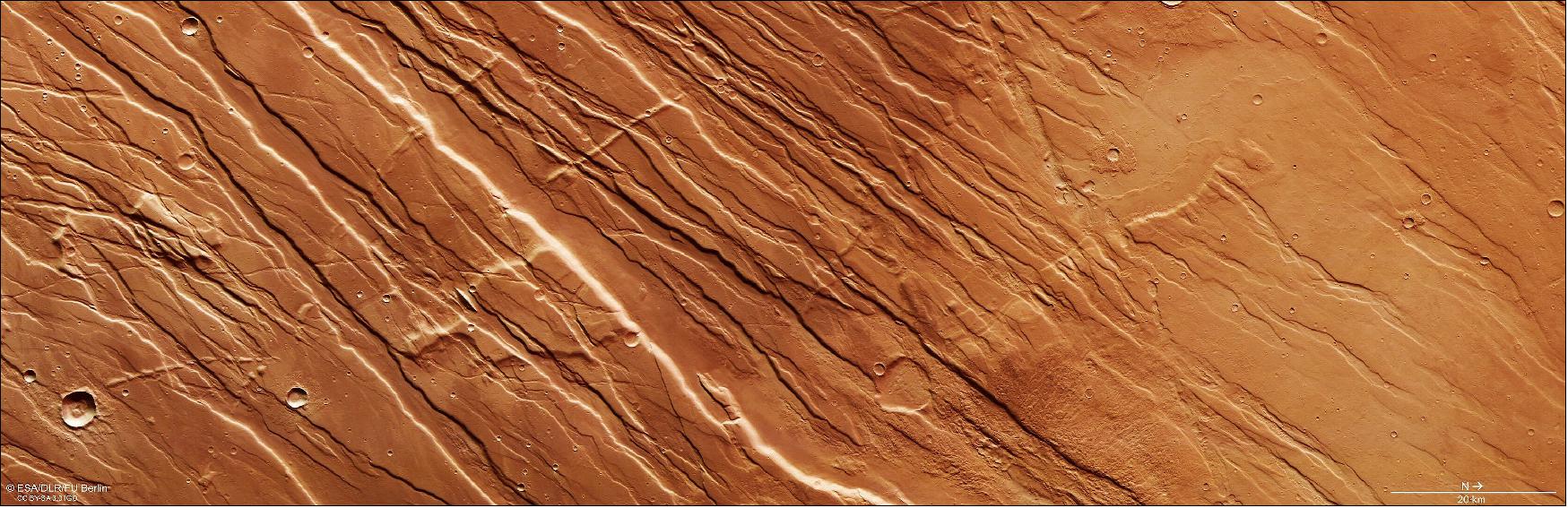 Figure 93: Located just northeast of the colossal Tharsis volcanic-tectonic province on Mars, the landscape shown in this image from ESA’s Mars Express is a mix of faults, elevated ground, deep valleys, and largely parallel ridges, extending both down into the surface and up above the martian crust. This is a portion of Tempe Fossae – a series of tectonic faults that cuts across Tempe Terra in Mars’ northern highlands. This region is a great example of terrain featuring two key martian features: grabens and horsts. In a way, these are opposites of one another – grabens are slices of ground that have dropped down between two roughly parallel faults, while horsts are ground that has been uplifted between faults. Both were created by tremendous volcanic and tectonic forces acting across the surface of Mars, which fractured the ground and manipulated it into new configurations. The surface to the right of the frame is smoother, created as lava flooded the region before cooling and solidifying, and some perpendicular slices across the predominantly parallel ridges can be seen to the left of the frame. As the nearby Tharsis province grew larger, it stretched and stressed the surrounding crust – and these features are evidence of a change in the direction of stress. This image comprises data gathered on 30 September 2019 during orbit 19913. The ground resolution is approximately 15 m/pixel and the images are centered at about 279°E/36°N. This image was created using data from the nadir and color channels of the High Resolution Stereo Camera (HRSC). The nadir channel is aligned perpendicular to the surface of Mars, as if looking straight down at the surface. North is to the right (image credit: ESA/DLR/FU Berlin, CC BY-SA 3.0 IGO)