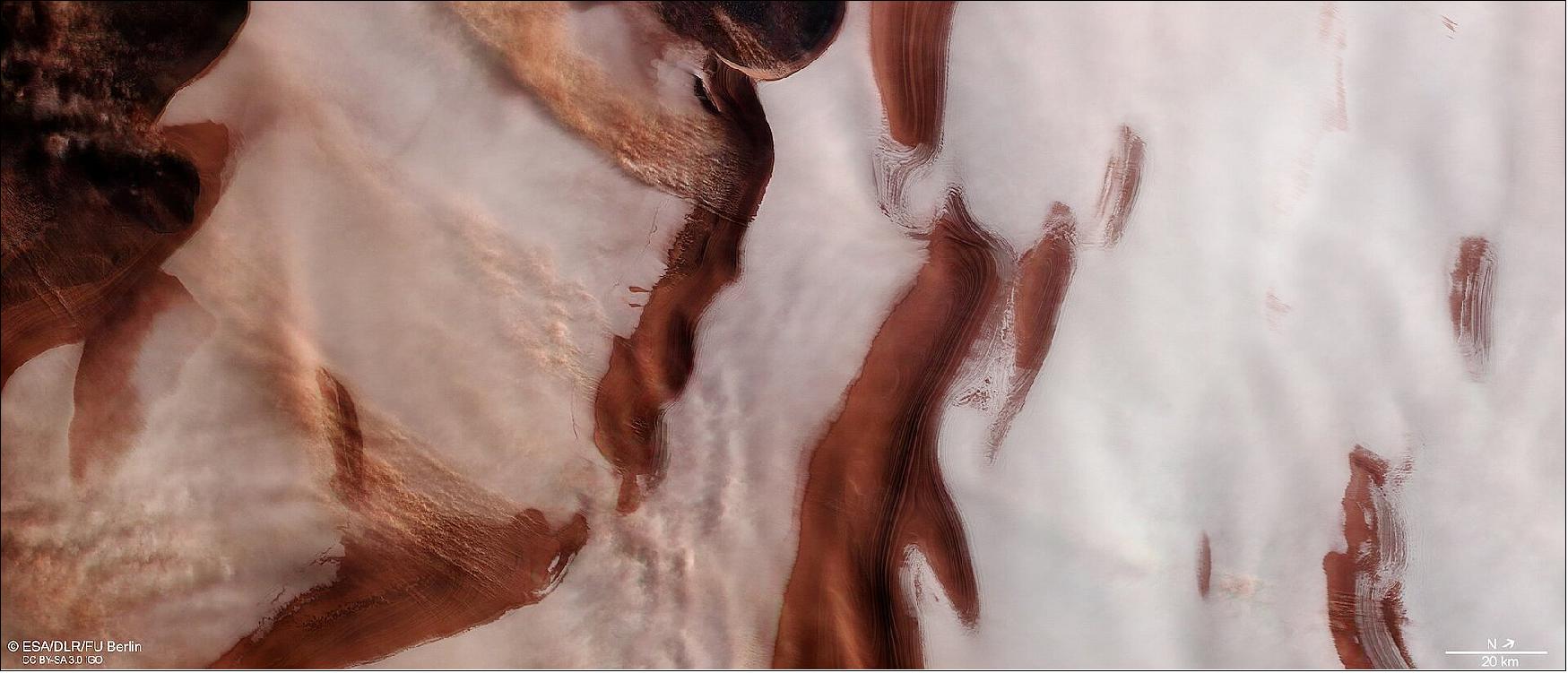 Figure 103: This image shows part of the ice cap sitting at Mars’ north pole, complete with bright swathes of ice, dark troughs and depressions, and signs of strong winds and stormy activity. The landscape here is a rippled mix of color. Dark red and ochre-hued troughs appear to cut through the icy white of the polar cap; these form part of a wider system of depressions that spiral outwards from the very center of the pole. Visible to the left of the frame are a few extended streams of clouds, aligned perpendicularly to a couple of the troughs. These are thought to be caused by small local storms that kick up dust into the martian atmosphere, eroding scarps and slopes as they do so and slowly changing the appearance of the troughs over time. - This image comprises data gathered on 16 November 2006 during orbit 3670. The ground resolution is approximately 15 m/pixel and the images are centered at about 244ºE/85ºN. This image was created using data from the nadir and color channels of the High Resolution Stereo Camera (HRSC). The nadir channel is aligned perpendicular to the surface of Mars, as if looking straight down at the surface. North is to the upper right (image credit: ESA/DLR/FU Berlin , CC BY-SA 3.0 IGO)